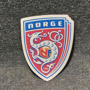 May include: A shield-shaped sticker featuring a red background with a white dragon illustration. The top blue banner displays the text NORGE in white. A circular emblem in the center contains the letters NF in red and blue, with the year 1902 below.