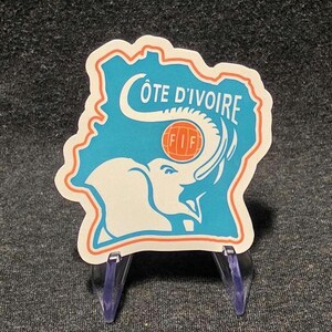 May include: A die-cut sticker shaped like the map of Côte d'Ivoire in light blue with an orange outline. It features a white elephant silhouette, an orange soccer ball with the text FIF, and the white text CÔTE D'IVOIRE across the top.