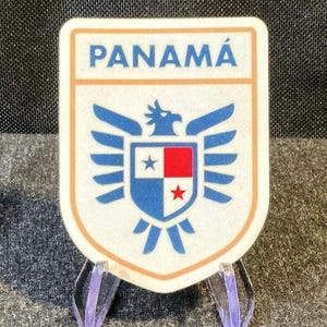 May include: A shield-shaped sticker featuring the word PANAMÁ in blue text at the top. Below the text is a blue eagle graphic centered over a shield divided into four quadrants of white, red, and blue, with a blue star and a red star.