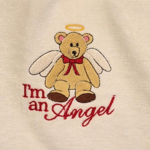 Puede incluir: Dise&ntilde;o bordado de un oso de peluche &aacute;ngel con halo y alas, con pajarita roja. El texto "I'm an Angel" est&aacute; bordado en rojo debajo. El fondo es de tela color crema.