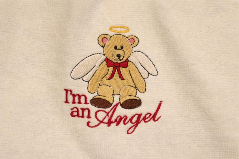 Puede incluir: Dise&ntilde;o bordado de un oso de peluche &aacute;ngel con halo y alas, con pajarita roja. El texto "I'm an Angel" est&aacute; bordado en rojo debajo. El fondo es de tela color crema.