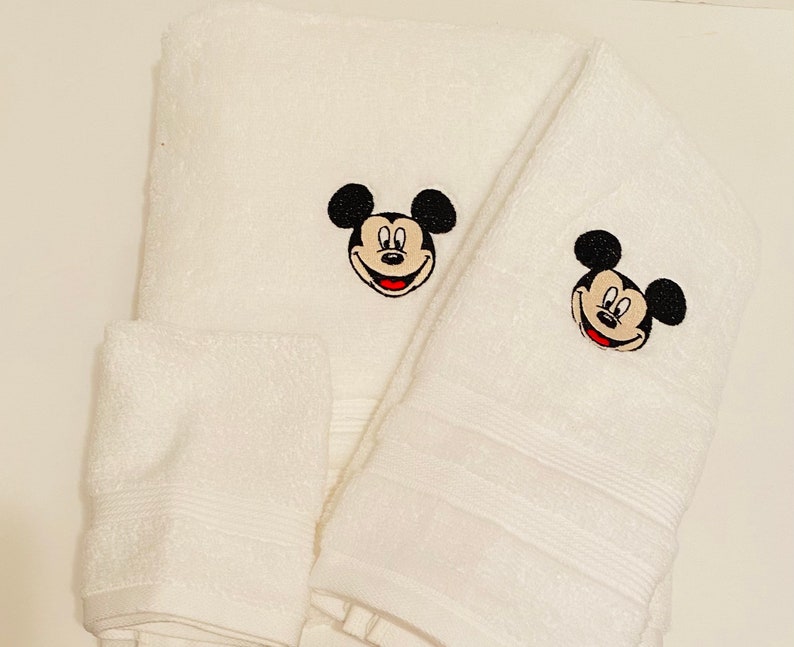 May include: A set of three white bath towels featuring embroidered Mickey Mouse faces. The towels are made from soft, absorbent cotton.