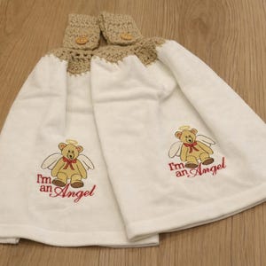 Puede incluir: Dos toallas de mano blancas con una parte superior de ganchillo y cierre de bot&oacute;n. Cada toalla presenta un oso de peluche bordado con alas de &aacute;ngel y el texto "I'm an Angel" en rojo. Las toallas se muestran sobre una superficie de madera.