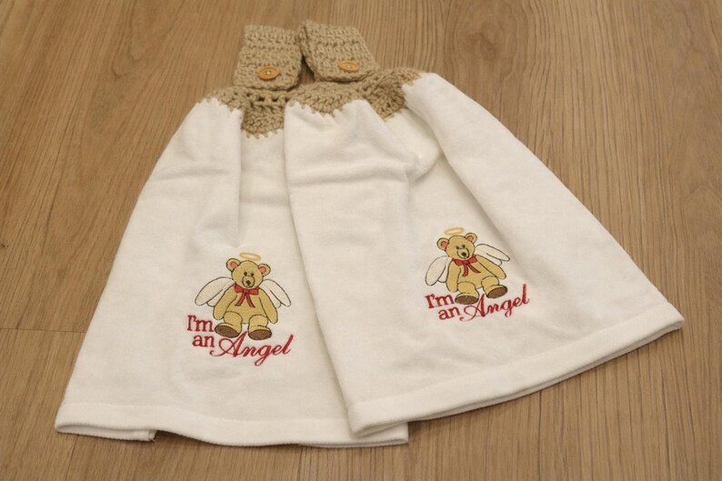 Puede incluir: Dos toallas de mano blancas con una parte superior de ganchillo y cierre de bot&oacute;n. Cada toalla presenta un oso de peluche bordado con alas de &aacute;ngel y el texto "I'm an Angel" en rojo. Las toallas se muestran sobre una superficie de madera.