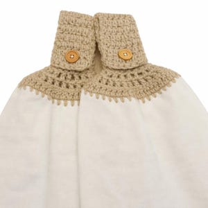 Puede incluir: Una toalla de cocina blanca con una parte superior de ganchillo beige y cierres de bot&oacute;n. La toalla tiene un borde festoneado decorativo. La parte superior de ganchillo est&aacute; sujeta con dos botones de madera.