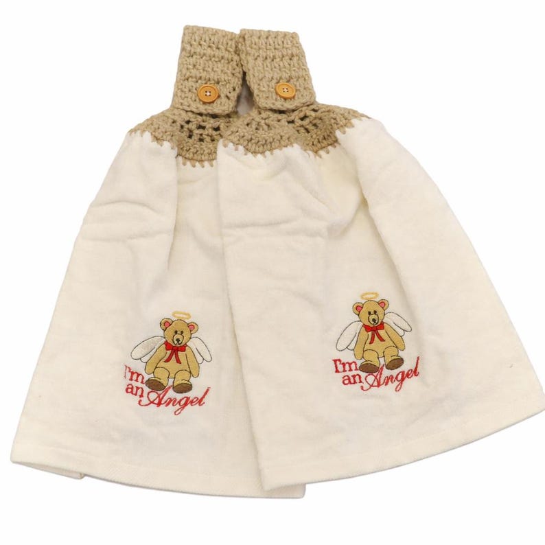 Puede incluir: Dos pa&ntilde;os de cocina de color crema con una parte superior de ganchillo beige y cierre de bot&oacute;n de madera. Cada toalla presenta un oso de peluche bordado con alas de &aacute;ngel, un halo y el texto "I'm an Angel" en rojo.