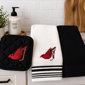 May include: A black quilted pot holder, a white kitchen towel with black stripes, and a solid black kitchen towel. Each item features an embroidered red cardinal bird perched on a brown branch. The set is displayed together in a kitchen setting.