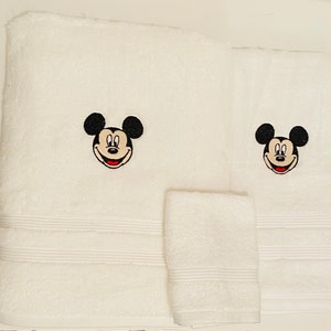 May include: White bath towels with embroidered Mickey Mouse faces. The set includes a hand towel, a bath towel, and a face cloth.
