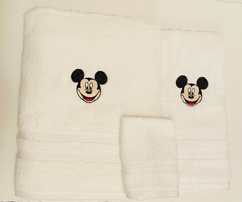 May include: White bath towels with embroidered Mickey Mouse faces. The set includes a hand towel, a bath towel, and a face cloth.
