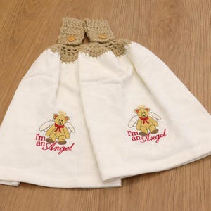 Puede incluir: Dos pa&ntilde;os de cocina blancos colgantes con una parte superior de ganchillo beige y cierre de bot&oacute;n. Cada toalla presenta un oso de peluche bordado con alas de &aacute;ngel y el texto "I'm an Angel" en rojo.