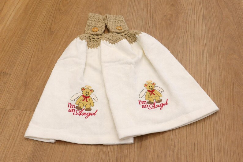 Puede incluir: Dos pa&ntilde;os de cocina blancos colgantes con una parte superior de ganchillo beige y cierre de bot&oacute;n. Cada toalla presenta un oso de peluche bordado con alas de &aacute;ngel y el texto "I'm an Angel" en rojo.