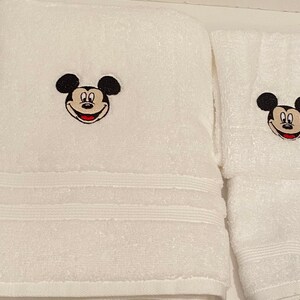 May include: White bath towels with embroidered Mickey Mouse faces. The larger towel has a single Mickey Mouse face, while the smaller towel has a Mickey Mouse face on each end.