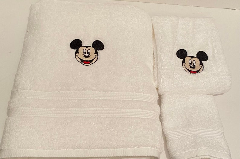 May include: White bath towels with embroidered Mickey Mouse faces. The larger towel has a single Mickey Mouse face, while the smaller towel has a Mickey Mouse face on each end.