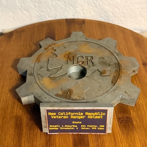 May include: A metallic gear-shaped display stand featuring an embossed bear silhouette, the letters NCR, and a star. A rectangular plaque is attached to the front, displaying the text: New California Republic Veteran Ranger Helmet, Stats, Weight: 1.5lbs/3kg, Hit Points: 100, Damage Threshold: 4, Value: 999 Caps.
