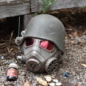 Op de afbeelding: Een verweerde, grijze helm met rood getinte ooglenzen, een gasmasker en een antenne. Daarnaast liggen bierdoppen, een kleine fles met een etiket en kleine blauwe kralen. Op een grindoppervlak.
