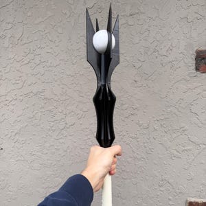 May include: A black staff featuring a pointed, multi-pronged crown structure at the top that cradles a smooth, white spherical orb. The staff has a long, cylindrical white handle extending downwards from the black, angular headpiece.