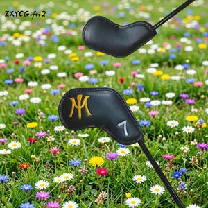 Puede incluir: Funda negra para la cabeza del palo de golf con un dise&ntilde;o dorado de la letra "M" y el n&uacute;mero "7" en blanco. La funda se encuentra sobre un fondo de un prado vibrante lleno de coloridas flores silvestres y hierba verde.