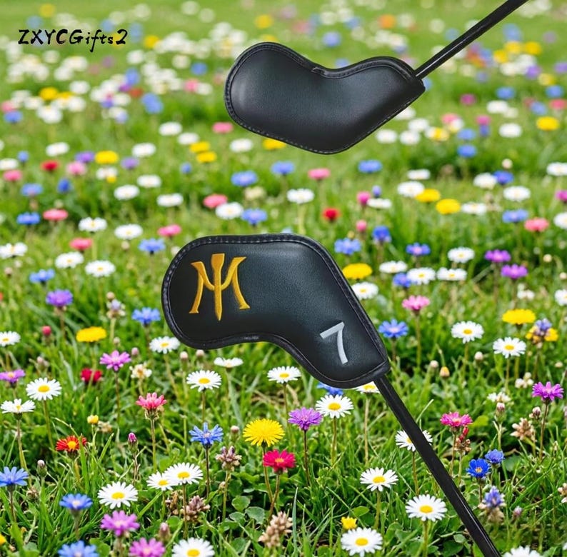Puede incluir: Funda negra para la cabeza del palo de golf con un dise&ntilde;o dorado de la letra "M" y el n&uacute;mero "7" en blanco. La funda se encuentra sobre un fondo de un prado vibrante lleno de coloridas flores silvestres y hierba verde.