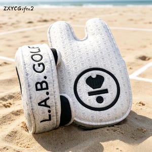 Könnte beinhalten: Zwei weiße Golfschlägerhauben mit schwarzen Akzenten und dem Text "L.A.B. GOLF". Die Hauben liegen auf Sand. Eine Haube hat ein rundes Emblem mit einer Krone und der Zahl "1/2". Die Hauben haben ein Muster aus kleinen Herzen.