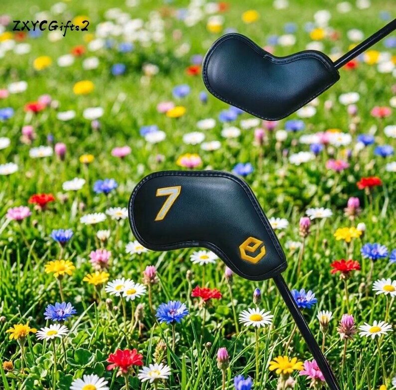 Puede incluir: Dos fundas negras para la cabeza de un palo de golf con n&uacute;meros y logotipos amarillos se muestran sobre un fondo de un prado vibrante lleno de coloridas flores silvestres. Las fundas est&aacute;n sobre ejes negros.