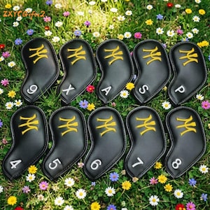 Puede incluir: Un juego de fundas negras para palos de golf con dise&ntilde;os bordados en dorado y n&uacute;meros y letras en blanco. Las fundas est&aacute;n dispuestas sobre un lecho de hierba verde y flores coloridas. El texto "ZXYCGifts2" es visible.