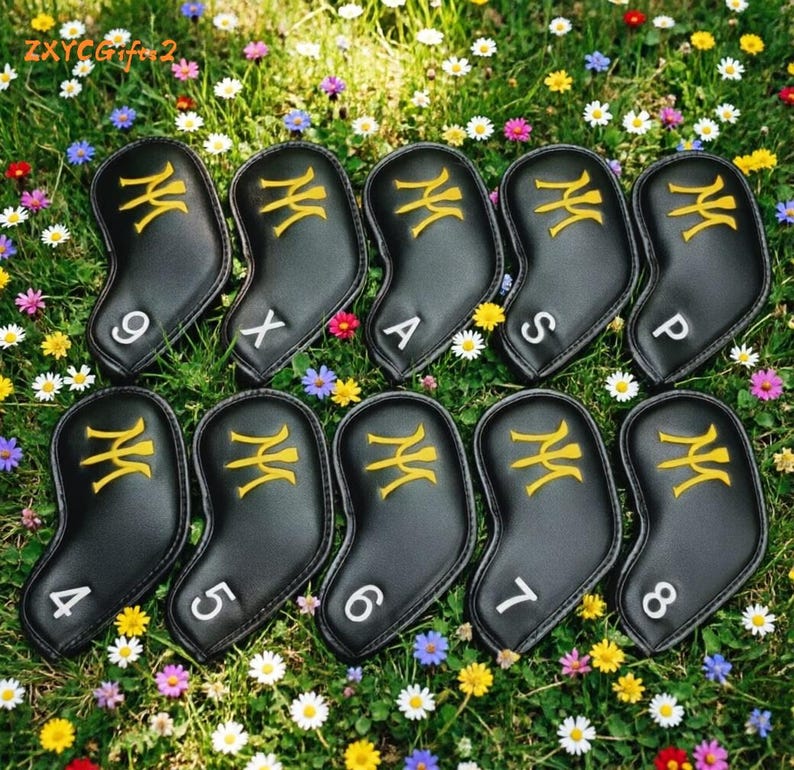 Puede incluir: Un juego de fundas negras para palos de golf con dise&ntilde;os bordados en dorado y n&uacute;meros y letras en blanco. Las fundas est&aacute;n dispuestas sobre un lecho de hierba verde y flores coloridas. El texto "ZXYCGifts2" es visible.