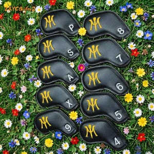 Puede incluir: Un conjunto de fundas negras para palos de golf con iniciales y n&uacute;meros dorados bordados. Las fundas est&aacute;n dispuestas sobre un lecho de hierba verde y flores coloridas, incluyendo margaritas.