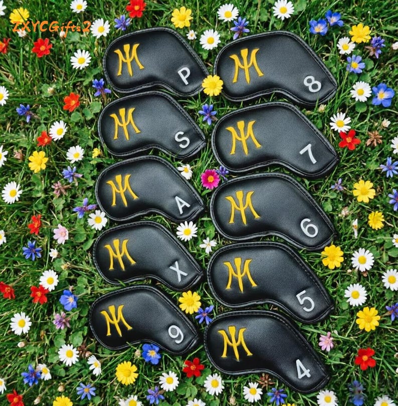 Puede incluir: Un conjunto de fundas negras para palos de golf con iniciales y n&uacute;meros dorados bordados. Las fundas est&aacute;n dispuestas sobre un lecho de hierba verde y flores coloridas, incluyendo margaritas.