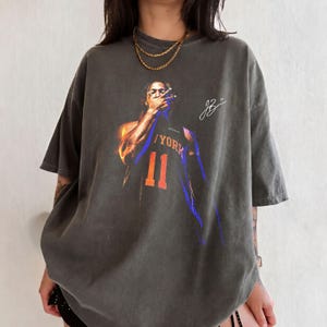 May include: A charcoal grey short-sleeve t-shirt featuring a graphic of a basketball player in a New York jersey with the number 11. The design includes a white signature and blue and orange accents. The shirt has a crew neckline and loose fit.
