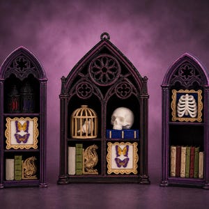 Op de afbeelding: Drie paarse, gotische displayplanken met spitsbogen, gevuld met miniatuur decoratieve voorwerpen zoals een witte menselijke schedel, een vogelkooi, ingelijste vlinders, een ribbenkastmodel en diverse kleine boeken op de planken.