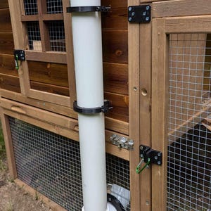 以下が含まれることがあります： 木製の鶏小屋の構造に垂直に取り付けられた白いPVCパイプの給餌器。円筒形のチューブの上部には丸いキャップ、下部には湾曲したエルボジョイントがあり、2つの黒いプラスチック製のクランプで木製のフレームに固定されています。