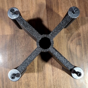 May include: A black, cross-shaped metal stand with a textured finish. The structure features a central circular opening and four cylindrical legs, each topped with a silver-toned circular plate and a small black screw fastener at the center.
