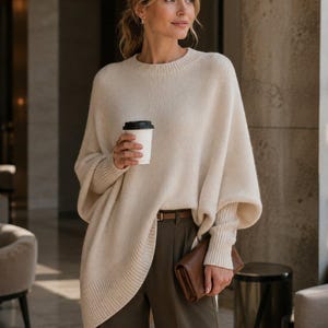 Op de afbeelding: Een crèmekleurige trui met wijde mouwen en een lange, asymmetrische zoom. De trui wordt gecombineerd met olijfgroene broek en een bruine leren riem. Een bruine clutch en een koffiekopje maken de look compleet.