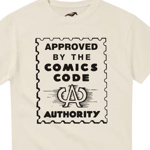 May include: Cream-colored t-shirt featuring a black graphic resembling a postage stamp. The stamp design includes the text "APPROVED BY THE COMICS CODE AUTHORITY" in bold, uppercase letters. A small horse logo is visible near the neckline.