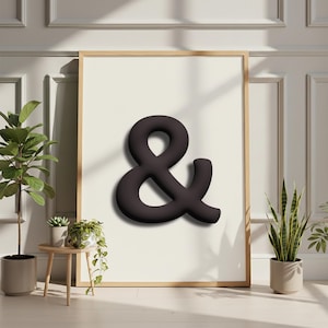 Puede incluir: Una impresión enmarcada con un gran símbolo de ampersand gris oscuro. El símbolo tiene una apariencia tridimensional y está centrado en un fondo crema. El marco es de madera clara, y la obra de arte se exhibe en una habitación con plantas.