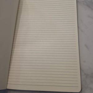 May include: An open notebook with cream-colored pages featuring thin, horizontal black lines. The paper has a smooth texture and rounded corners on the right side. The notebook is positioned to show a single page spread for writing or drawing.