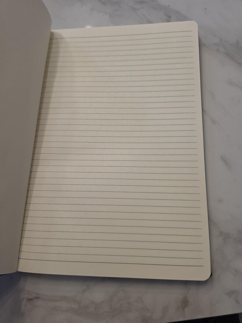 May include: An open notebook with cream-colored pages featuring thin, horizontal black lines. The paper has a smooth texture and rounded corners on the right side. The notebook is positioned to show a single page spread for writing or drawing.