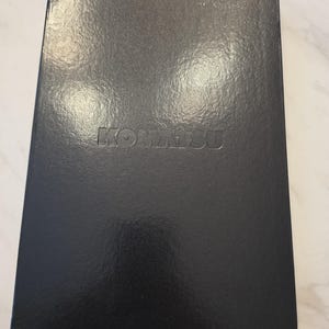 May include: A rectangular black notebook with a glossy finish and a smooth texture. The word KOMATSU is embossed in capital letters in the center of the front cover. The notebook has sharp, clean edges and a solid, dark, uniform appearance.