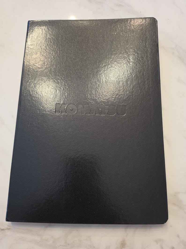 May include: A rectangular black notebook with a glossy finish and a smooth texture. The word KOMATSU is embossed in capital letters in the center of the front cover. The notebook has sharp, clean edges and a solid, dark, uniform appearance.