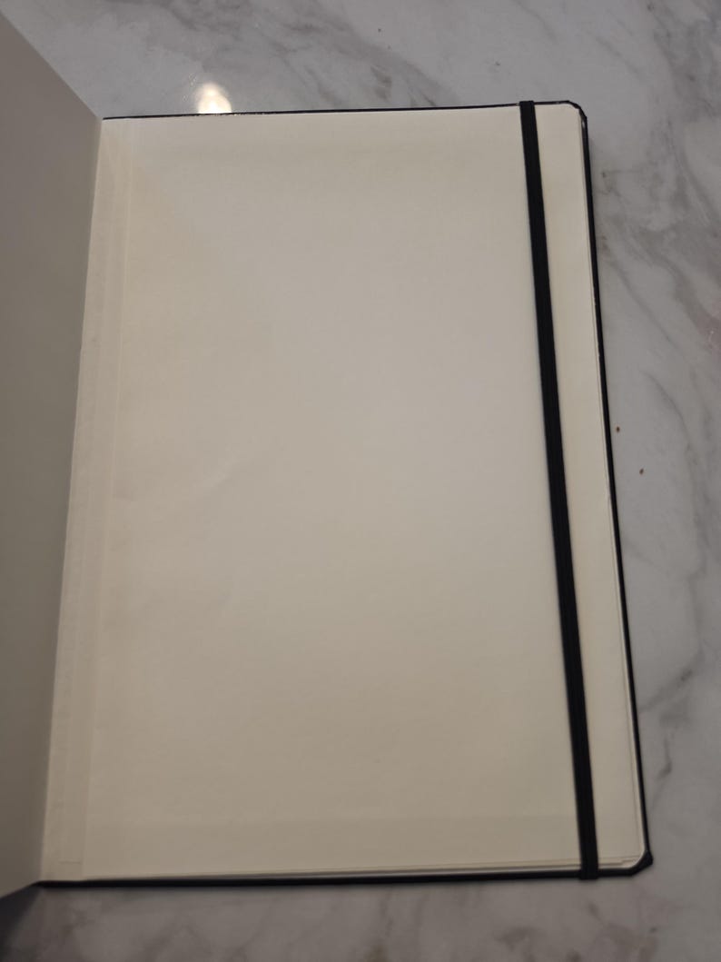 May include: An open notebook with blank, off-white pages and a black elastic band closure. The notebook features a sturdy black cover and is positioned to show the interior paper surface, which is smooth and unlined, providing a clean writing area.
