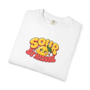 May include: A white t-shirt featuring a graphic of a grumpy yellow lemon character with a green leaf. The text "SOUR BY NATURE" is written in bold, bubbly, orange and red letters surrounding the lemon, accented by small star shapes.