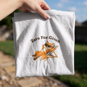 May include: A white t-shirt featuring a graphic of an orange fox wearing black sunglasses, lounging in a blue and white striped beach chair. Above the fox, the text reads Zero Fox Given in brown, sans-serif, arched lettering.