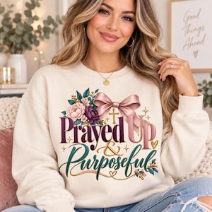 Puede incluir: Una sudadera de cuello redondo color crema con un diseño gráfico que incluye un lazo rosa, detalles florales, una pequeña cruz dorada y el texto "Prayed Up & Purposeful" en letras cursivas de color morado oscuro y verde azulado.