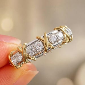 May include: A two-tone ring with a silver band and gold accents. The ring features multiple clear gemstones set in a row, with gold X-shaped designs wrapping around the band. The ring is held up by a person's fingers.