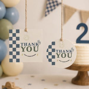 May include: Two hanging paper gift tags, one square and one circular, featuring a blue checkered pattern, a smiley face, a peace sign, and the text THANK YOU LOVE CHRISTOPHER with lightning bolt icons, suspended by thin twine strings against a neutral background.
