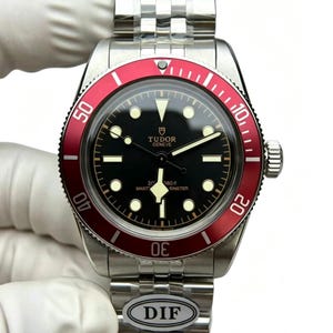 May include: A stainless steel wristwatch with a black dial and a red bezel. The watch features luminous hour markers and hands, and a stainless steel bracelet. The watch face displays the brand name "Tudor Geneve".