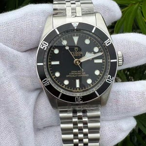 May include: A stainless steel wristwatch with a black dial and a black rotating bezel. The watch face features white hour markers and hands, with the brand name "TUDOR GENEVE" visible. The watch band is a silver-colored metal link bracelet.