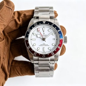 Könnte beinhalten: Eine Edelstahl-Armbanduhr mit weißem Zifferblatt, rot-blauer Lünette und silbernem Armband. Die Uhr hat eine Datumsanzeige und den Text "GMT Chronometer Officially Certified". Die Uhr wird von einer braunen behandschuhten Hand gehalten.