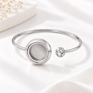 May include: A silver-toned open cuff bracelet featuring a large circular setting with a smooth, light grey centre and a smaller, separate circular setting adorned with a single clear, faceted crystal stone at the opposite end of the band.