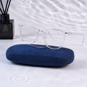 Può includere: Occhiali con montatura in plastica trasparente dalla classica forma arrotondata e dettagli in metallo argentato sulle cerniere, appoggiati su una custodia rigida in tessuto denim blu scuro con cuciture arancioni visibili.